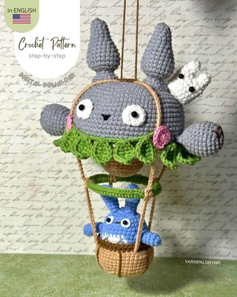 May include: A crocheted gray character with a white bunny and a blue character in a basket, suspended by strings. The gray character has large eyes and a green leafy collar. The image includes the text "Crochet Pattern" and "Digital Download".