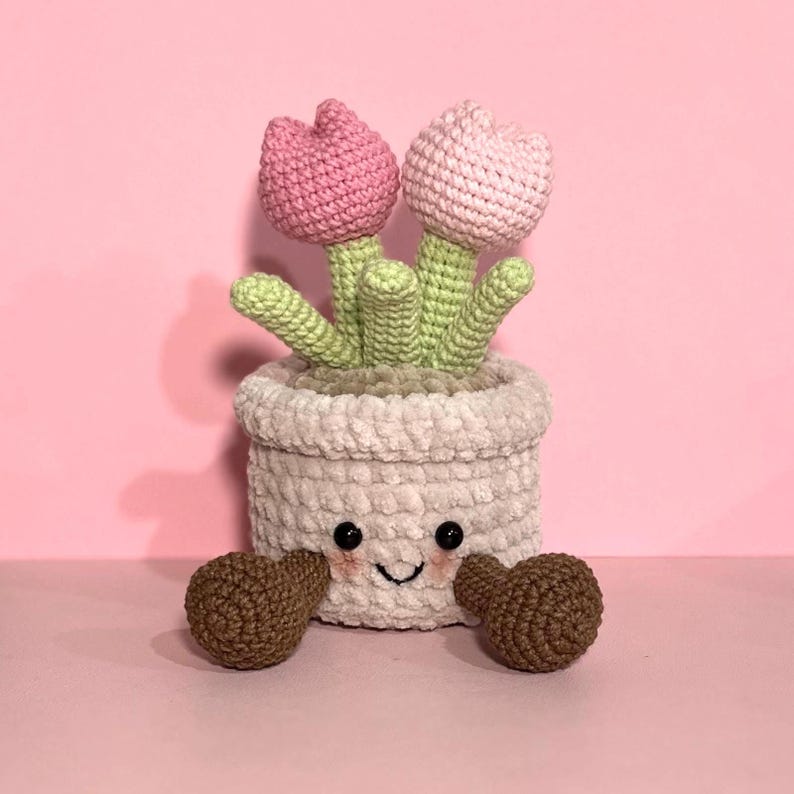 May include: A crocheted plush toy shaped like a potted plant with two pink tulips. The pot has a smiling face and brown crocheted arms.