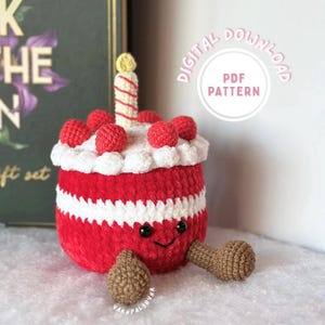 May include: A crocheted red and white birthday cake with a smiling face, topped with red spheres and a candle. The cake has brown legs and the text "DIGITAL DOWNLOAD PDF PATTERN".