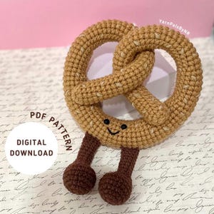 Puede incluir: Un pretzel de amigurumi tejido a crochet con una cara sonriente y dos piernas marrones. El pretzel es marrón y tiene una superficie texturizada. El texto "PDF PATTERN" y "DIGITAL DOWNLOAD" es visible en la imagen.