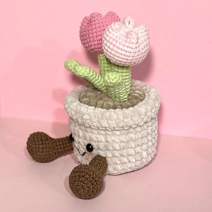 May include: A crocheted flower pot with two pink flowers and green leaves. The pot has brown legs and a cute face.