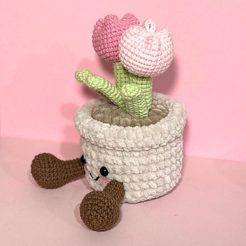 May include: A crocheted flower pot with two pink flowers and green leaves. The pot has brown legs and a cute face.