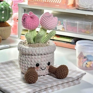 May include: A crocheted plush toy featuring two pink tulips emerging from a light beige pot. The pot has a smiling face with black eyes and rosy cheeks, and two brown crocheted legs extending outward from the base of the pot.