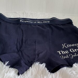 May include: Dark blue boxer shorts with white text reading "Property of Mrs Dow" on the waistband. The right leg features "Kenny The Groom 2nd July 2022" in white script. The shorts are on a white fluffy surface.