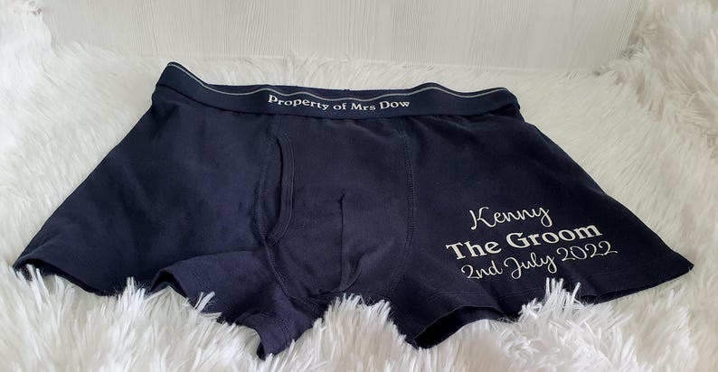May include: Dark blue boxer shorts with white text reading "Property of Mrs Dow" on the waistband. The right leg features "Kenny The Groom 2nd July 2022" in white script. The shorts are on a white fluffy surface.