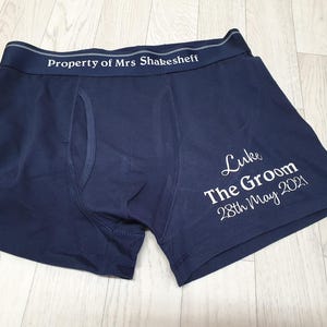 May include: Navy blue boxer shorts with white text reading "Property of Mrs Shakesheff" on the waistband. The shorts also feature the text "Luke The Groom 28th May 2021" on the leg. The shorts are made of a soft material.