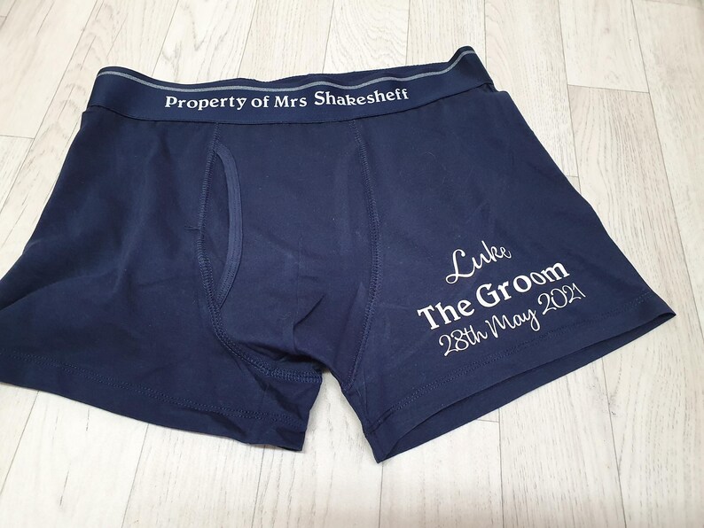May include: Navy blue boxer shorts with white text reading "Property of Mrs Shakesheff" on the waistband. The shorts also feature the text "Luke The Groom 28th May 2021" on the leg. The shorts are made of a soft material.
