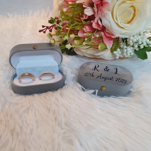 May include: Two gold wedding rings in a grey velvet ring box, next to a matching box with the inscription "R & L 27th August 2022". A bouquet of pink and cream flowers is in the background.