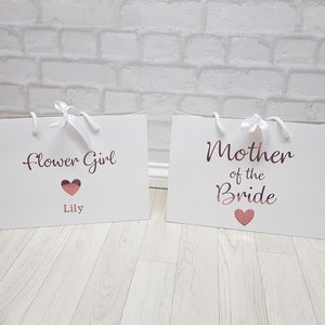 May include: Two white gift bags with rose gold lettering. The bag on the left says "Flower Girl" with a heart and the name "Lily". The bag on the right says "Mother of the Bride" with a heart.
