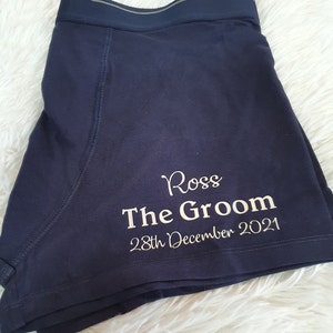 May include: Navy blue boxer shorts with the name "Ross", the text "The Groom", and the date "28th December 2021" printed in white. The shorts have a dark blue waistband with a gray stripe.