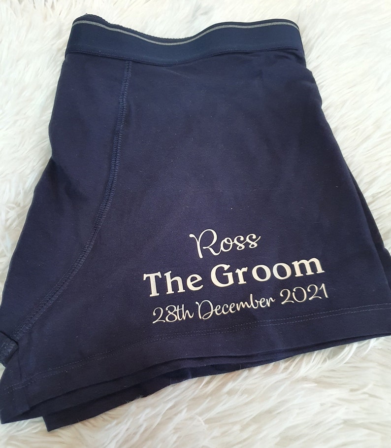 May include: Navy blue boxer shorts with the name "Ross", the text "The Groom", and the date "28th December 2021" printed in white. The shorts have a dark blue waistband with a gray stripe.