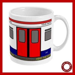 Op de afbeelding: Een witte keramische mok met een rood, wit en blauw ontwerp van een Londense metrodeur. De mok heeft de tekst "Mind the Gap" en "1973" op de bodem. De tekst "Your Name" is op de mok gedrukt.