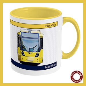 May include: A white ceramic mug with a yellow rim and handle. The mug features a graphic of a yellow tram with the text "Piccadilly" and "Metrolink" on the front. The mug is a product of Mind The Gap Graphic Design.