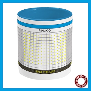 Puede incluir: Taza de cerámica blanca con borde azul y un gráfico en blanco y negro de una señal de estación de metro de Londres. El letrero dice "PIMLICO" y "MIND THE GAP".