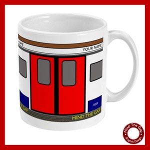 Op de afbeelding: Een witte keramische mok met een afbeelding van een rode Londense metrodeur met de tekst "Mind the Gap" en "2009" in blauw. De mok heeft een bruine streep en de tekst "Your Name" in zwart.