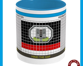 May include: A white ceramic mug with a blue interior and a design featuring a castle emblem. The mug has a grid pattern with black, red, and white squares. Text includes "Mind the Gap" and the names of two locations.