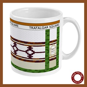 Puede incluir: Taza de cerámica blanca con un gráfico de un diseño de azulejos de la estación de metro de Londres. El diseño de los azulejos presenta un patrón marrón y blanco con las palabras "Trafalgar Square" en texto naranja. La taza tiene un asa verde.