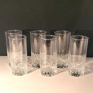 May include: Six clear, tall drinking glasses. The glasses have a faceted base and vertical lines on the sides. The glasses are made of clear glass and are arranged in a group.