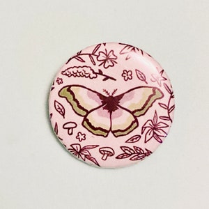 May include: A pink button with a white butterfly and floral design. The butterfly has brown and green wings with a pink body. The floral design includes leaves, flowers, and mushrooms.