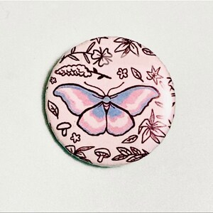May include: A pink button with a butterfly design. The butterfly has blue and pink wings and is surrounded by floral and mushroom illustrations.