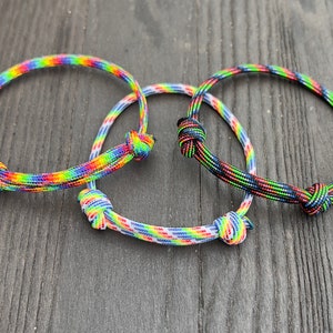 May include: Three colourful paracord bracelets. Each bracelet features a rainbow pattern with a knot closure. The bracelets are in shades of red, orange, yellow, green, blue, and purple. The bracelets are on a wooden surface.