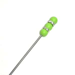 May include: A silver-colored hat pin with a decorative top. The top features three green beads, each encircled with a ring of clear rhinestones. The pin is designed to secure hats or other accessories.