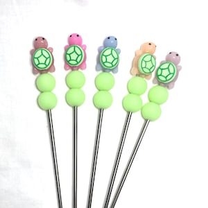 May include: Set of six cocktail picks with a whimsical turtle design. Each pick features a stainless steel stem, two light green spheres, and a colorful turtle topper. The turtles are in shades of pink, blue, and peach.