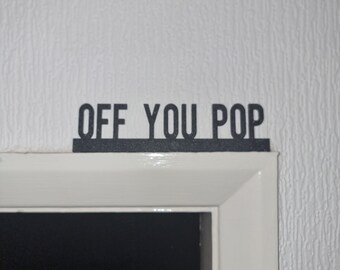 May include: Black sign with the words "OFF YOU POP" in a bold, sans-serif font. The sign is mounted above a white door frame, against a textured white wall. The sign is a decorative item.