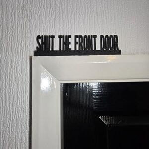 Pode incluir: Uma placa preta com o texto "Shut the front door" montada acima de uma moldura de porta branca.
