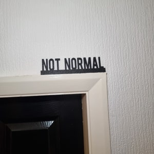 May include: A white door frame with the words "NOT NORMAL" printed in black letters above the door.