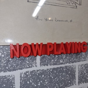 以下が含まれることがあります： 赤いプラスチック製の看板に、太字の3D文字で「NOW PLAYING」と書かれています。看板はレンガの壁に取り付けられています。