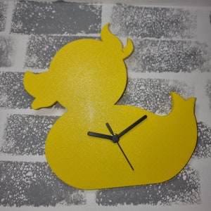 May include: Yellow rubber ducky shaped clock with black hands on a gray brick wall background.