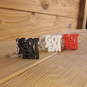 May include: Three keychains with the words "GOT EM" in black, white, and red. The keychains are made of a textured material and have a silver metal ring.