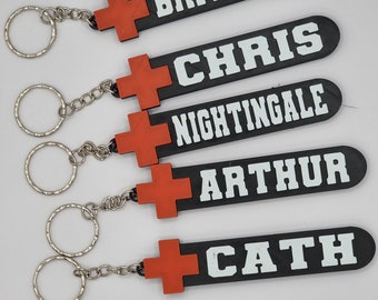 Puede incluir: Cinco llaveros negros con cruces rojas y texto blanco. Los nombres en los llaveros son Brittain, Chris, Nightingale, Arthur y Cath.
