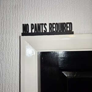 Könnte beinhalten: Ein schwarzes Schild mit dem Text "NO PANTS REQUIRED" über einem weißen Türrahmen.