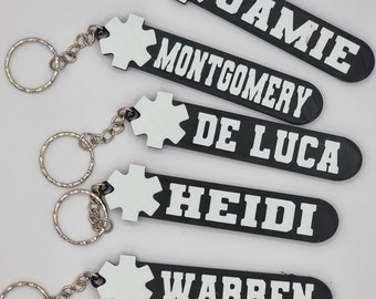 Puede incluir: Cinco llaveros negros con dijes en forma de estrella blanca y texto blanco. Los llaveros tienen los nombres "Jamie", "Montgomery", "De Luca", "Heidi" y "Warren".