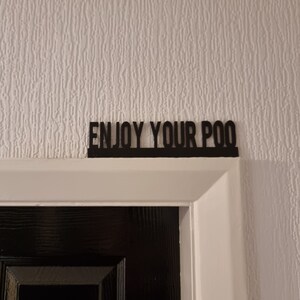 Może przedstawiać: Czarny znak z tekstem "ENJOY YOUR POO" zamontowany nad czarnymi drzwiami.