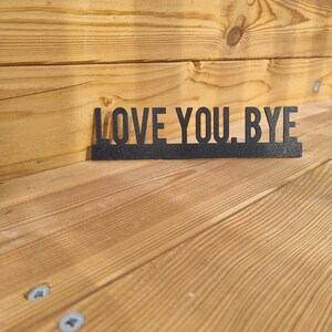 Op de afbeelding: Een zwart houten bord met de tekst "LOVE YOU. BYE" op een lichtbruin houten oppervlak.