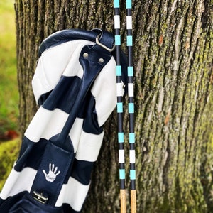 May include: A white and navy blue striped golf bag with a leather handle and a handprint logo. Two wooden golf clubs with black and turquoise stripes are leaning against a tree trunk.