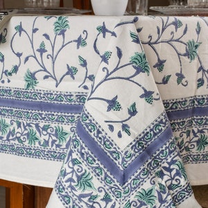 May include: White tablecloth with a floral pattern in shades of blue and green. The design features stylized flowers and vines, with a border of intricate patterns. The tablecloth is draped over a wooden table, showcasing the fabric's texture and design.