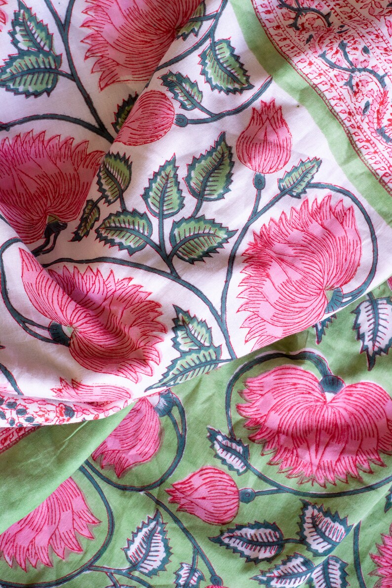May include: Close-up of floral fabric with a repeating pattern of pink flowers and green leaves on a white and green background. The fabric is folded, showing the detailed design.