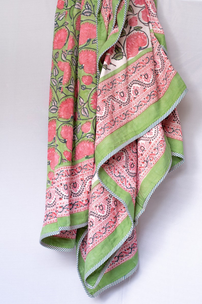 May include: A floral patterned textile featuring large pink flowers and green leaves on a light green background. The border is a contrasting green with a white and blue striped trim. The textile is folded, revealing a pink and white patterned interior.