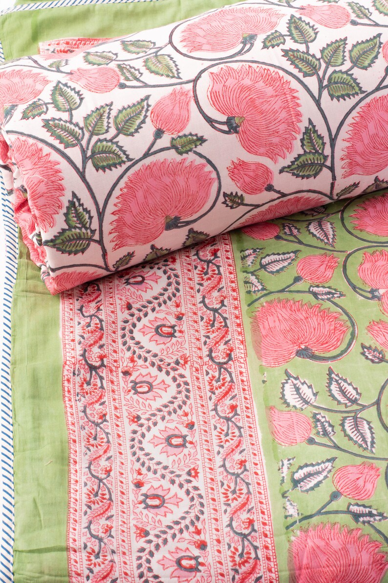 May include: A folded textile featuring a floral pattern in pink and green. The design includes large pink flowers with green leaves on a white background. A green textile with a floral border is also visible.