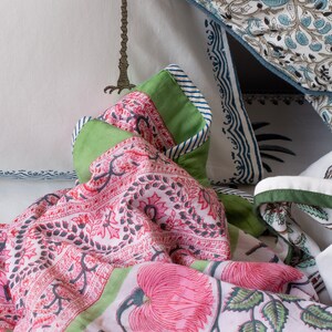 May include: A close-up of bedding with floral patterns in pink, green, and white. The quilt features large pink flowers and green leaves. Pillows with blue and white patterns are visible in the background. The quilt has a green border.