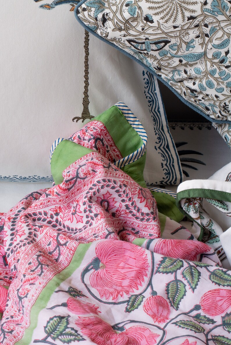 May include: A close-up of bedding with floral patterns in pink, green, and white. The quilt features large pink flowers and green leaves. Pillows with blue and white patterns are visible in the background. The quilt has a green border.