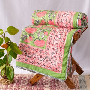 May include: A green and pink floral patterned quilt, partially rolled, rests on a wooden stool. The quilt features large pink flowers and a decorative border. A matching rug with a burgundy pattern is visible below.
