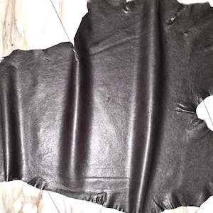 Könnte beinhalten: Ein Stück dunkelgraues Leder mit strukturierter Oberfläche. Das Leder hat eine natürliche, unregelmäßige Form mit zerrissenen Kanten. Das Material scheint flexibel zu sein und hat einen leichten Glanz, was darauf hindeutet, dass es behandelt wurde.
