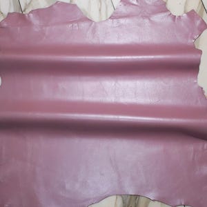 Könnte beinhalten: Ein Blatt aus mauvefarbenem Leder mit glatter, glänzender Oberfläche. Das Leder ist gefaltet, wodurch horizontale Falten in der Mitte entstehen. Die Kanten des Leders sind uneben, mit einer natürlichen, organischen Form.