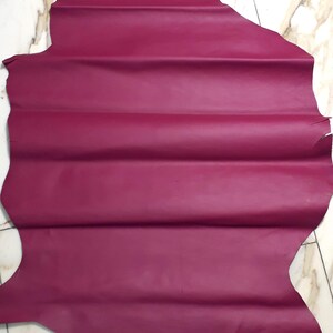 May include: A large, irregularly shaped piece of magenta leather. The leather has a smooth, matte finish and is folded in several places. The background is a light-coloured surface with a marble-like pattern.