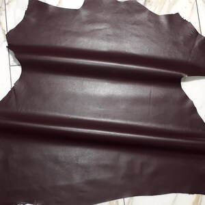 May include: A large, irregularly shaped piece of dark brown leather. The leather has a smooth, slightly glossy surface with natural folds and creases. The edges are raw and unfinished, revealing the texture of the material. Suitable for crafting.